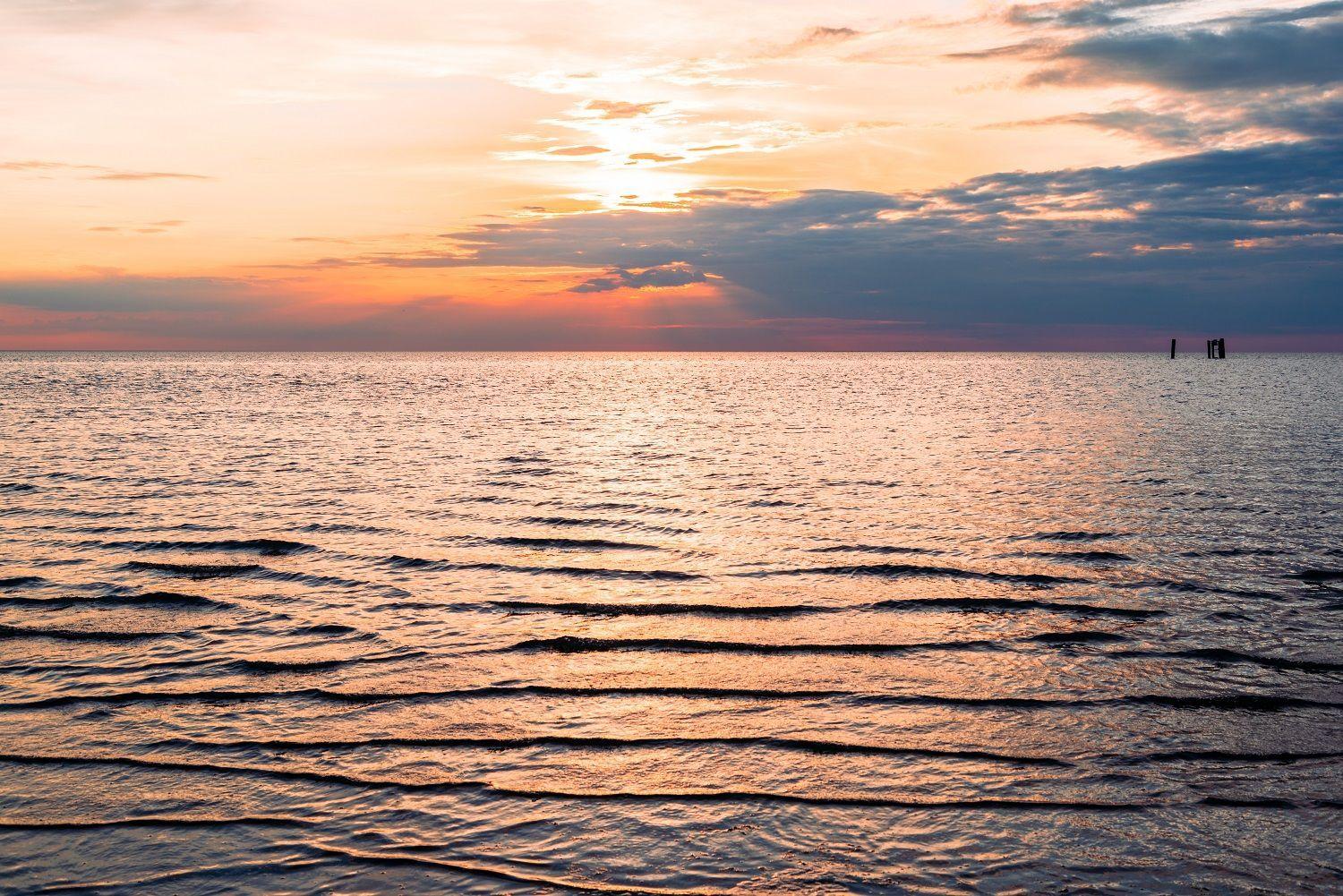 Sonnenuntergang über ruhigem Wasser mit Wellen und zwei Pfosten im Hintergrund.