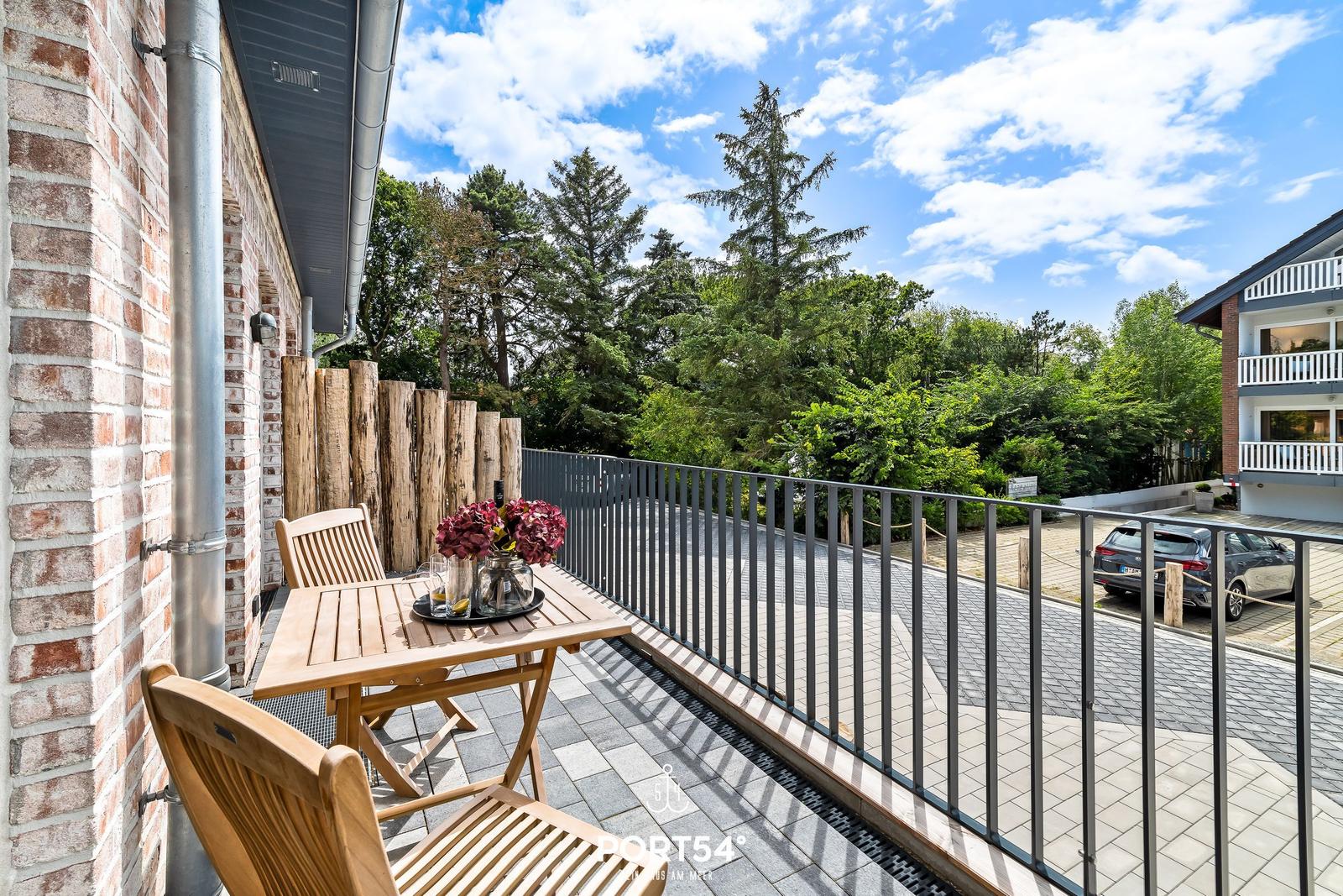 Terrasse mit Holztisch, Stühlen und Blumen. Blick auf grüne Bäume und einen gepflasterten Weg.