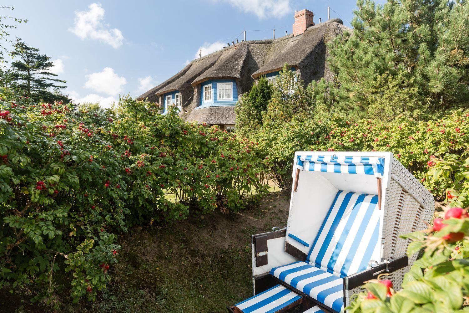 Ein Strandkorb steht im Garten vor einem strohgedeckten Haus mit blauen Fenstern.
