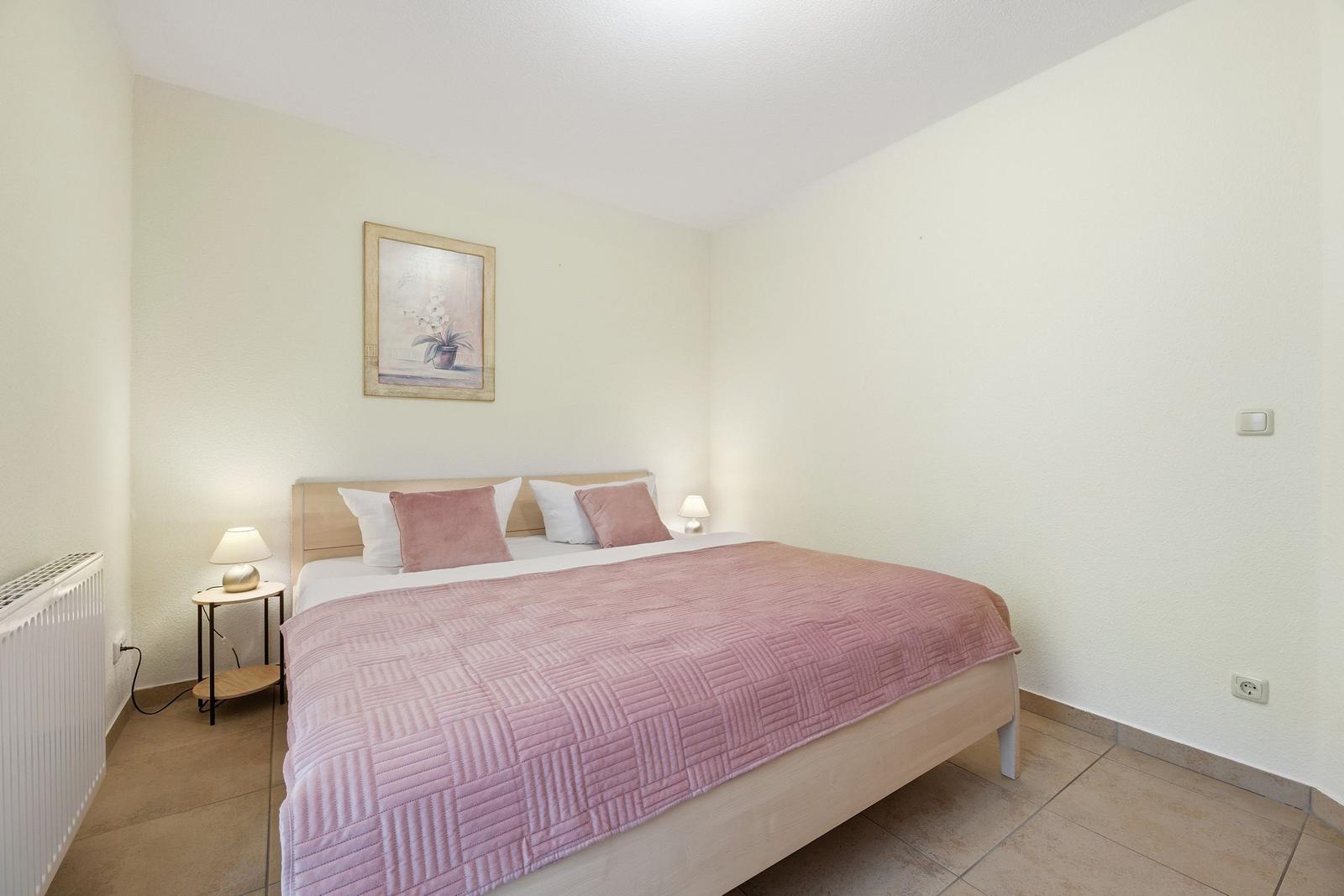 Schlafzimmer mit Bett, rosa Decke und zwei Nachttischen mit Lampen.
