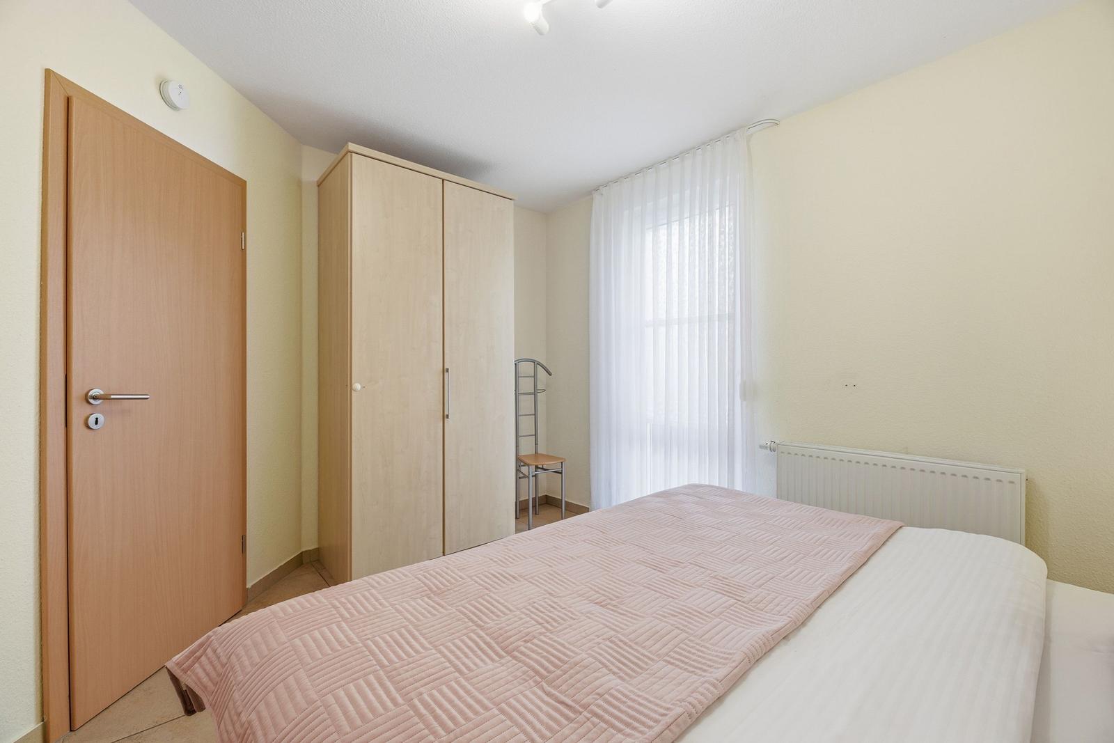 Schlafzimmer mit Bett, Kleiderschrank und Fenster mit weißen Vorhängen.