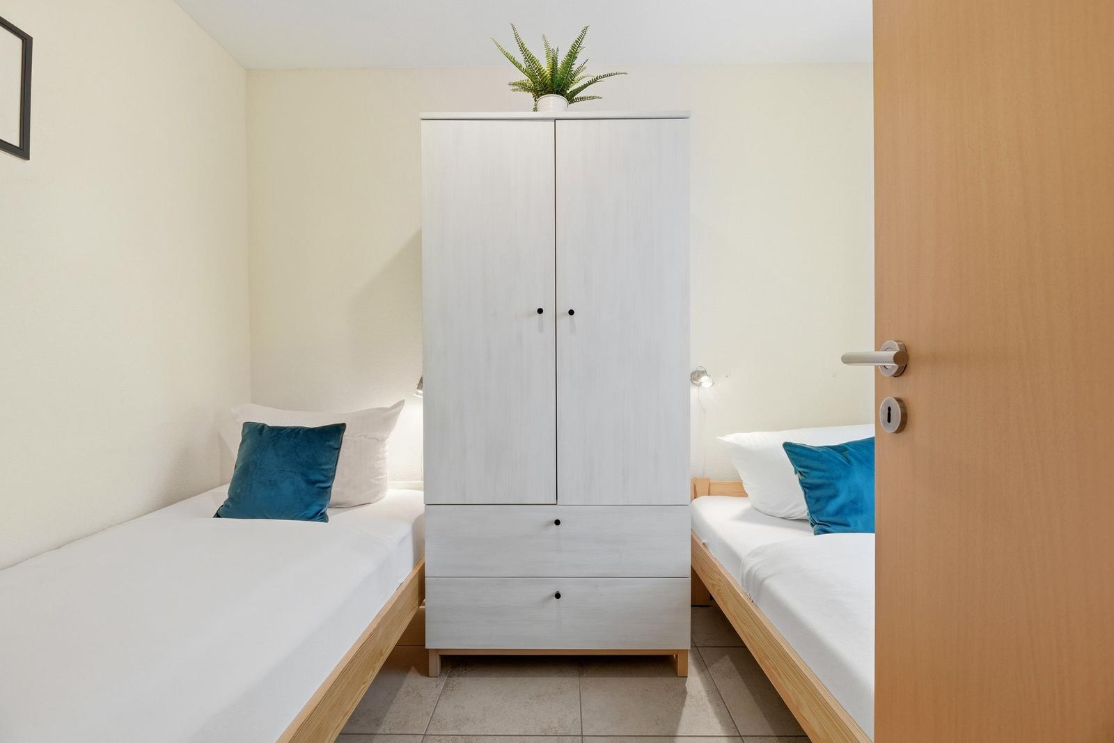 Zimmer mit zwei Betten, weißem Schrank und Pflanze.