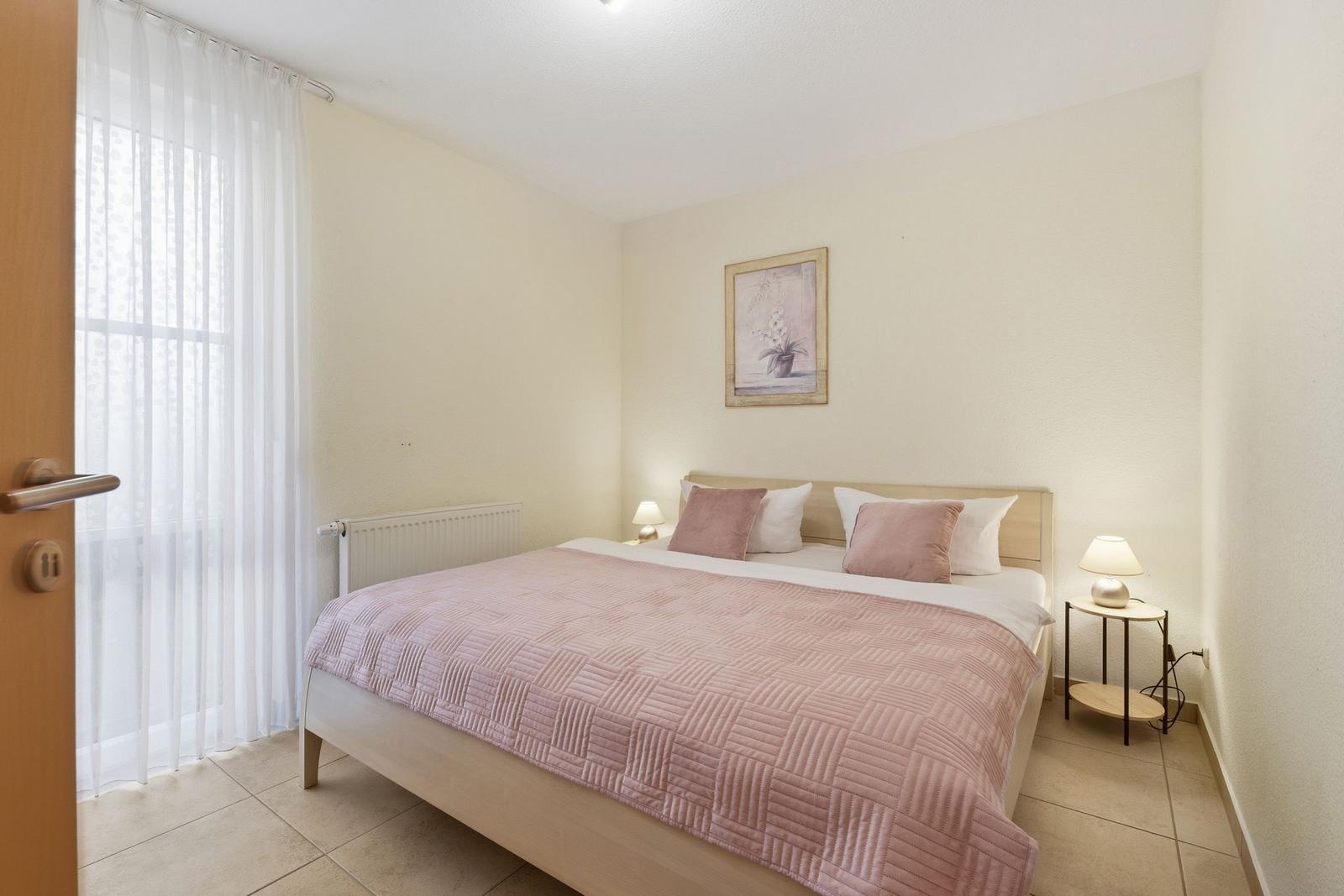 Schlafzimmer mit Bett, Nachttisch und Fenster mit weißen Vorhängen.