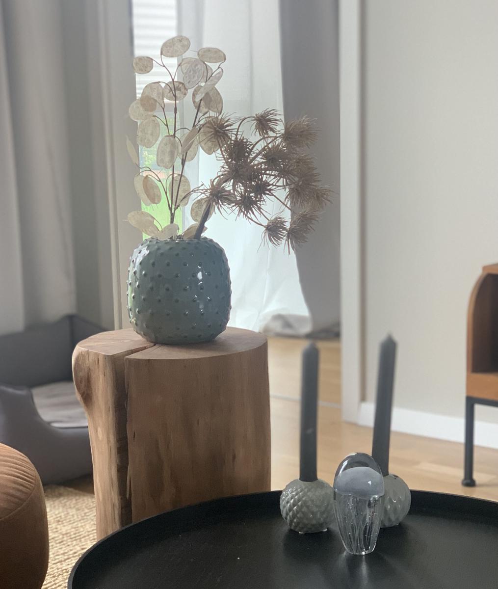 Ein grauer Vase mit Trockenpflanzen steht auf einem Holzblock. Vorherige Tisch mit Kerzen und Glasobjekten.