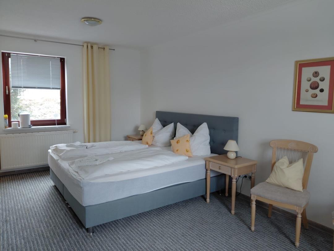 Schlafzimmer mit Doppelbett, Nachttisch, Stuhl und Fenster mit Jalousie.