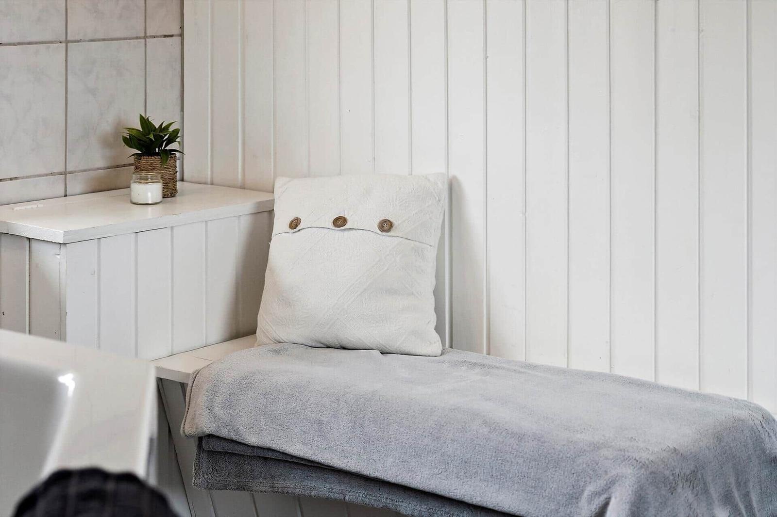 Badezimmerbank mit Kissen und Decke, Wand mit vertikalen Paneelen, Pflanze und Kerze auf Oberfläche.