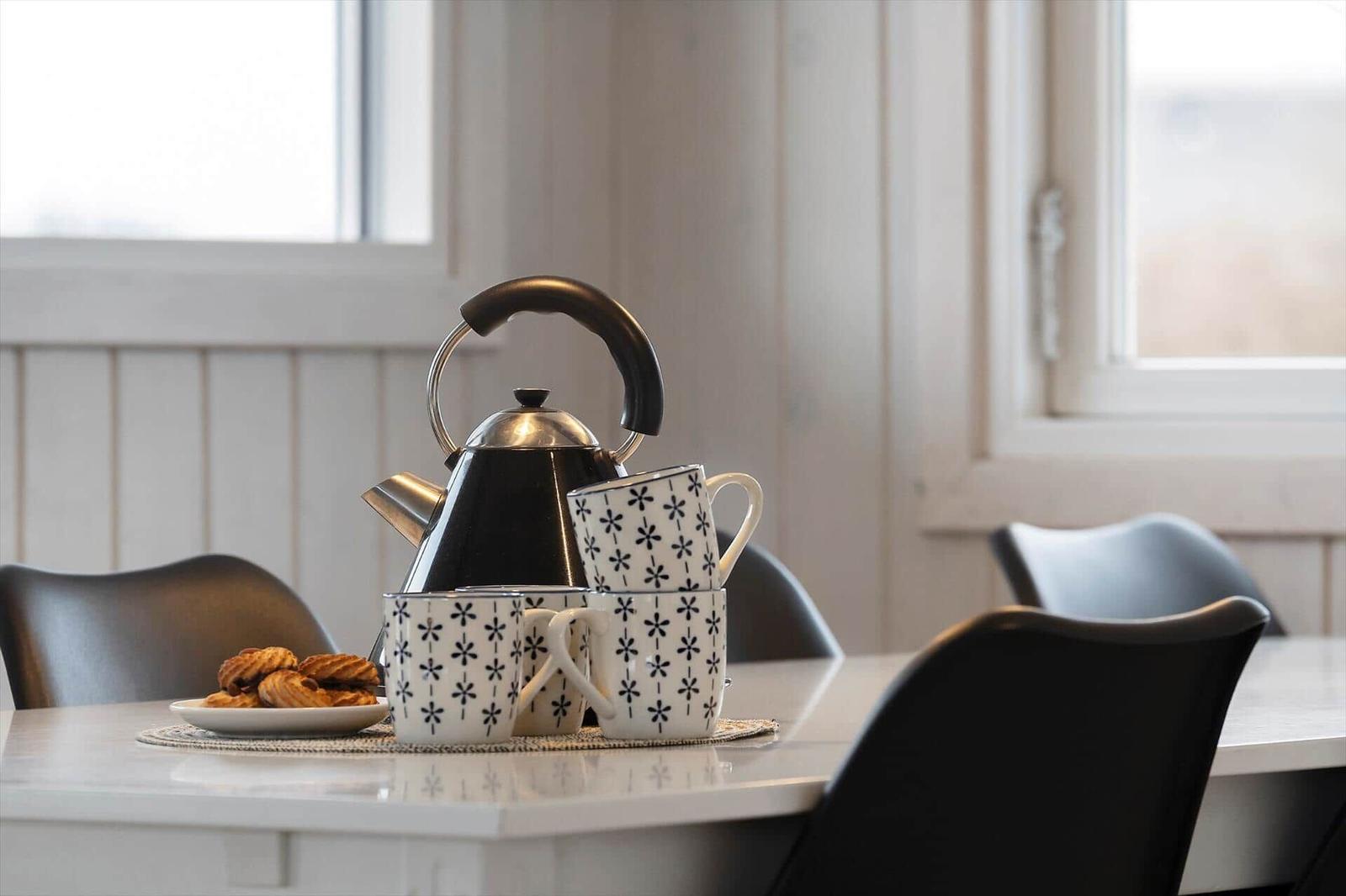 Tisch mit Kanne, Tassen und Keksen vor Fenstern.