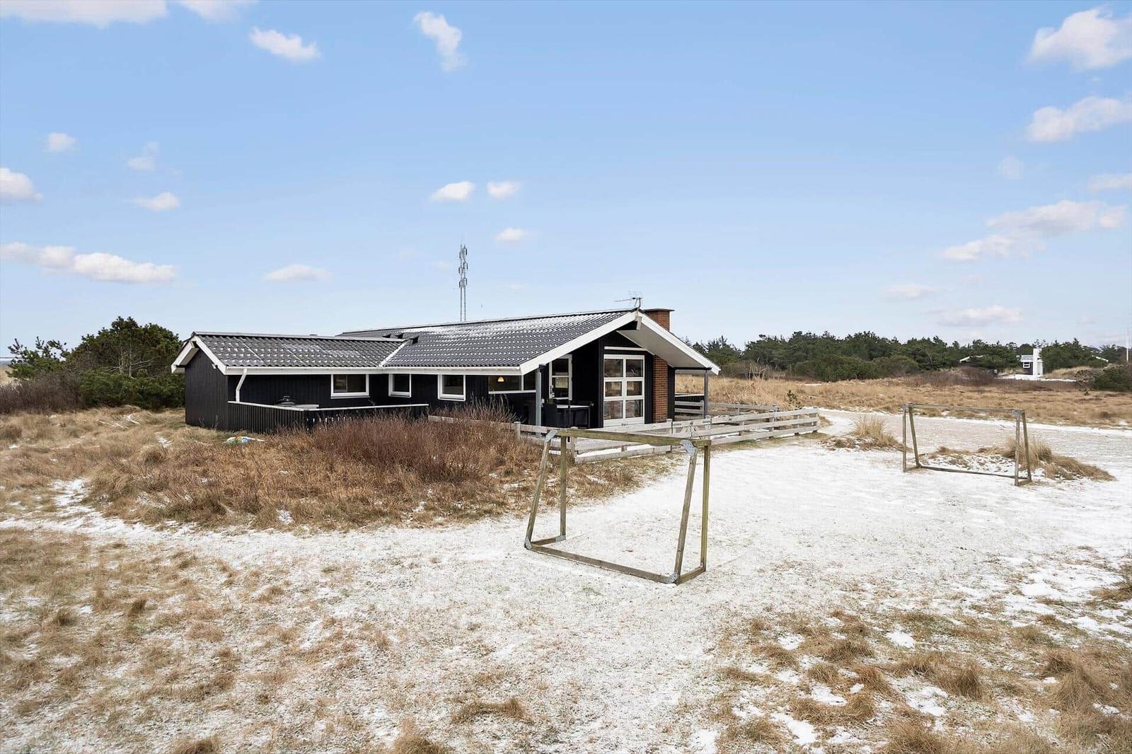 Ein schlichtes Haus mit Dach und Fenstern steht auf einer sandigen Fläche mit einem Fußballtor.