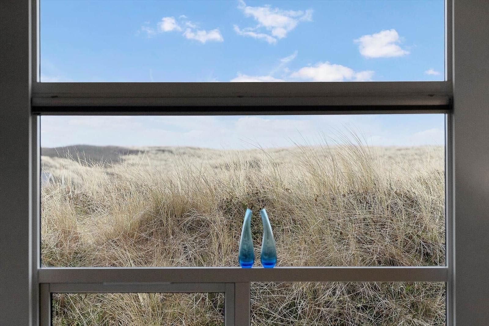 Durch das Fenster blickt man auf eine Wiese mit hohem Gras und zwei blauen Vasen.