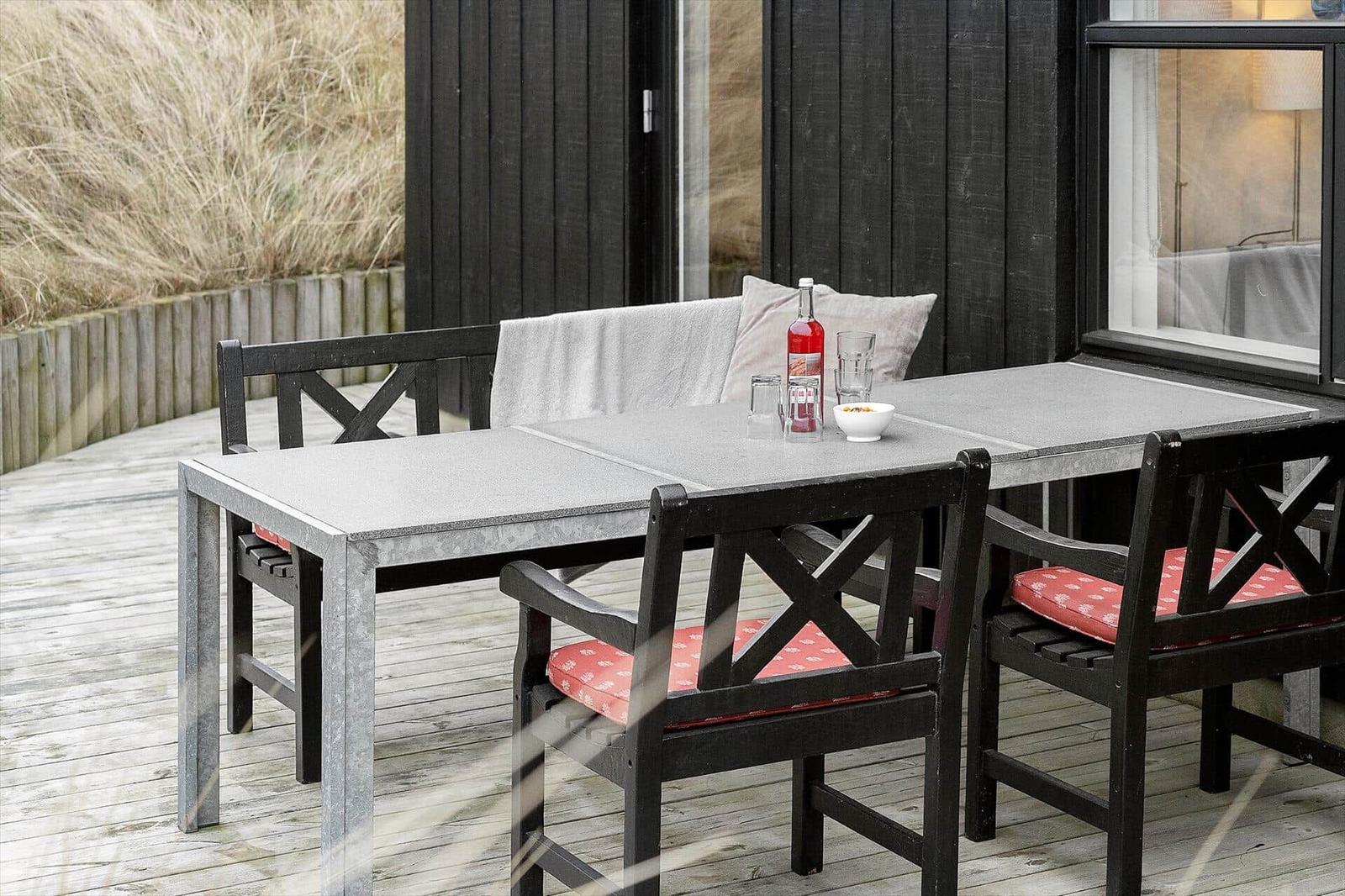Terrasse mit Tisch und Stühlen, rote Sitzkissen, Getränke und Blick auf Gras.