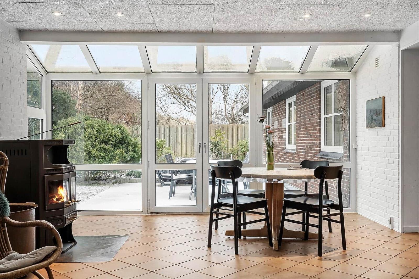 Sitzbereich mit Holztisch, Stühlen und Kaminofen. Große Fensterfront mit Blick auf Garten.