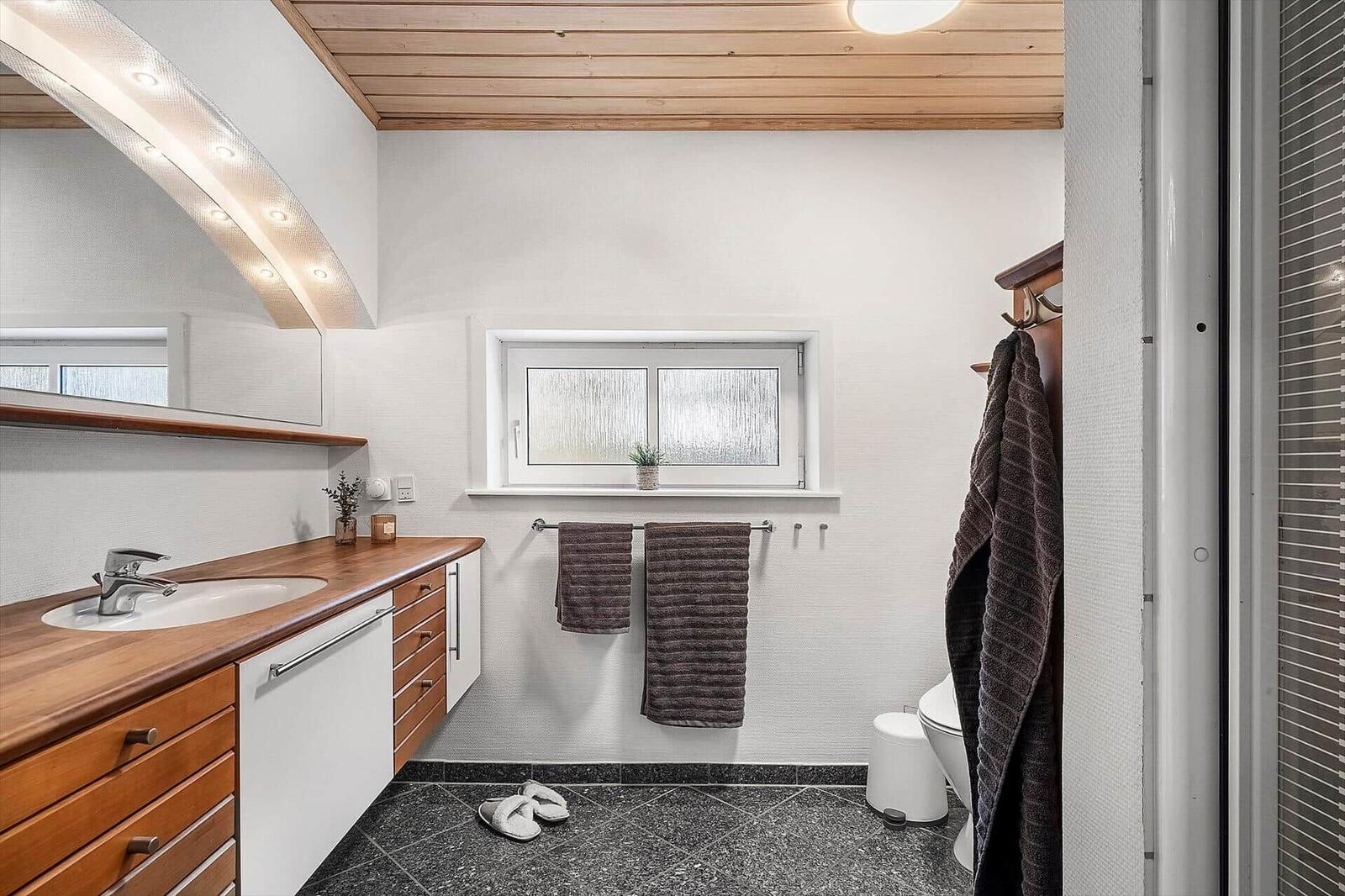 Badezimmer mit Holzoberfläche, Spiegel und Fenster. Handtuchhalter und Toilette sichtbar.