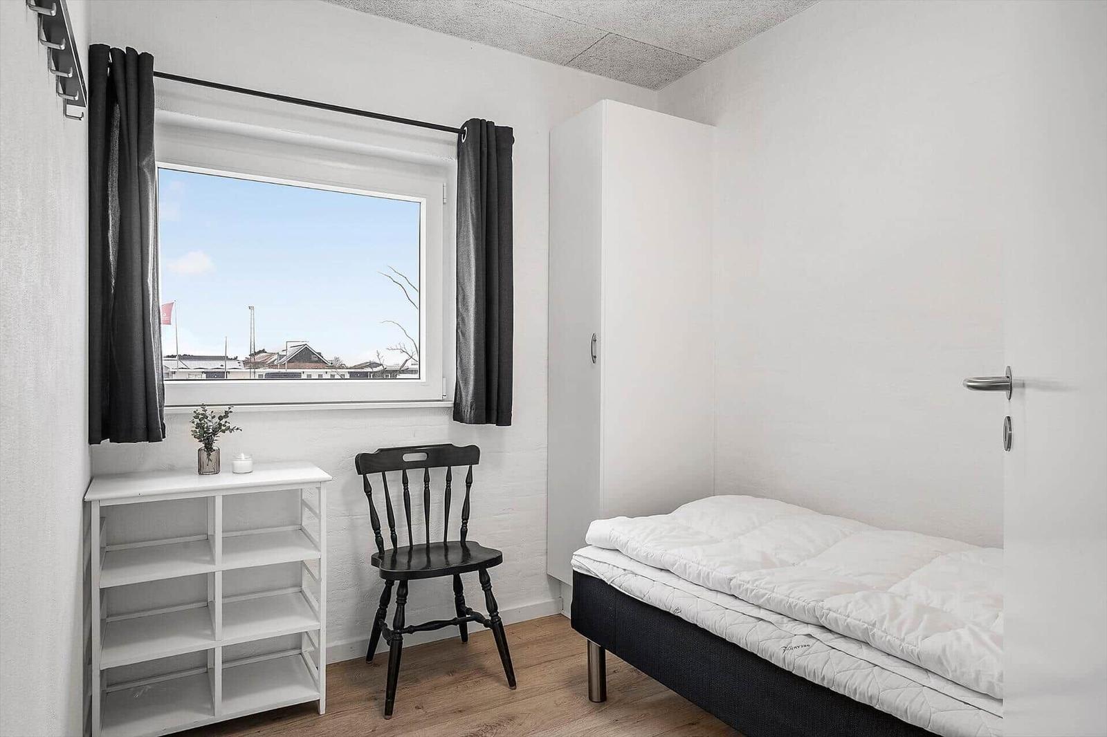 Schlafzimmer mit Bett, Stuhl und Fenster. Weißer Schrank und Holzboden.