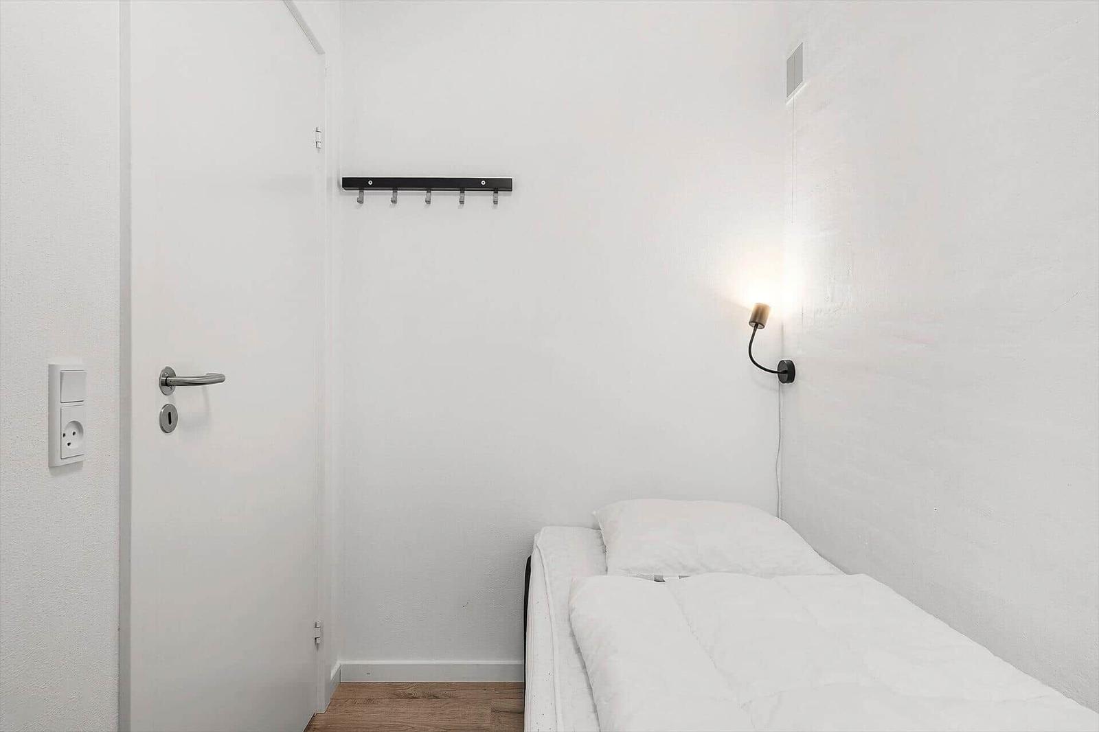 Ein schlichtes Schlafzimmer mit weißem Bett, Wandlampe und Kleiderhaken.