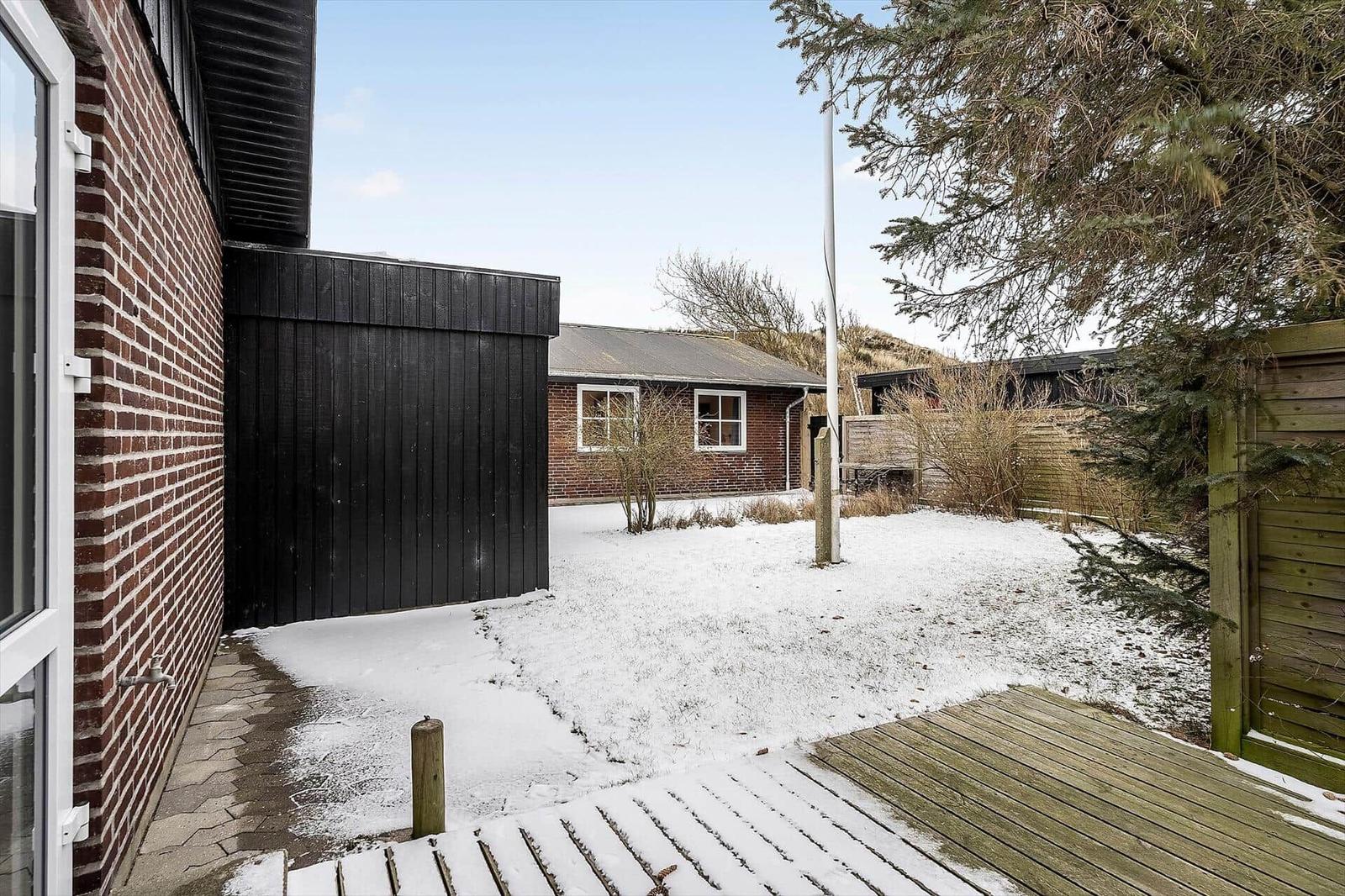 Schneebedeckter Garten mit Holzterrasse und rotem Backsteinhaus im Hintergrund.
