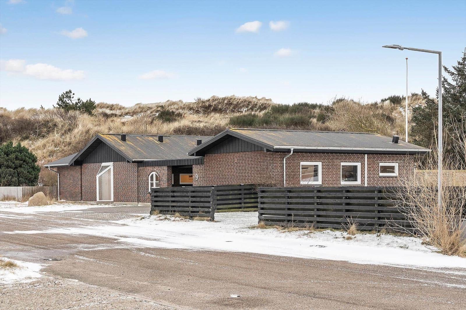 Ein zweistöckiges Haus mit Ziegelfassade und dunklem Dach, umgeben von Dünen und einem Holzzaun.