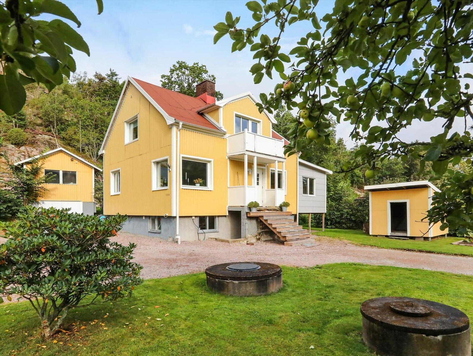 Gelbes Haus mit Balkon und Garten. Zwei kleinere Häuser und Bäume sind sichtbar.