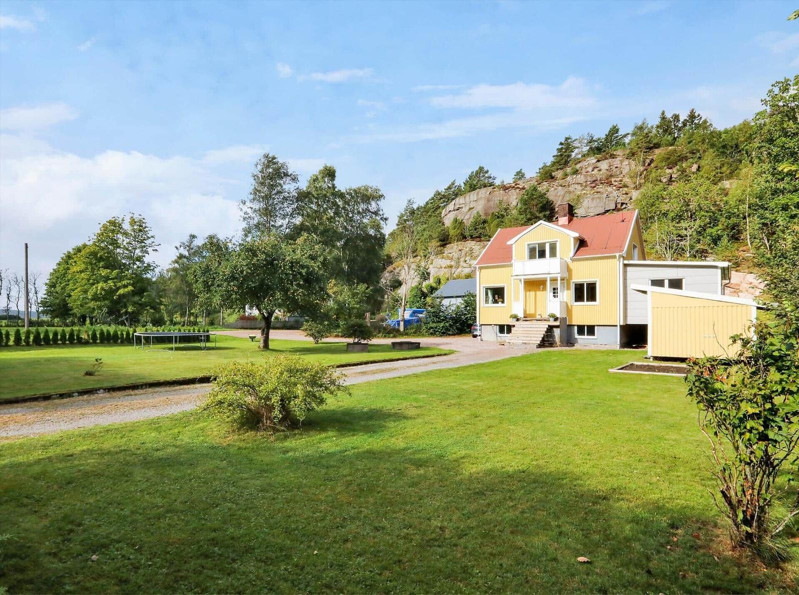 Ein gelbes Haus mit rotem Dach, umgeben von Gras und Bäumen. Ein Trampolin ist im Garten.