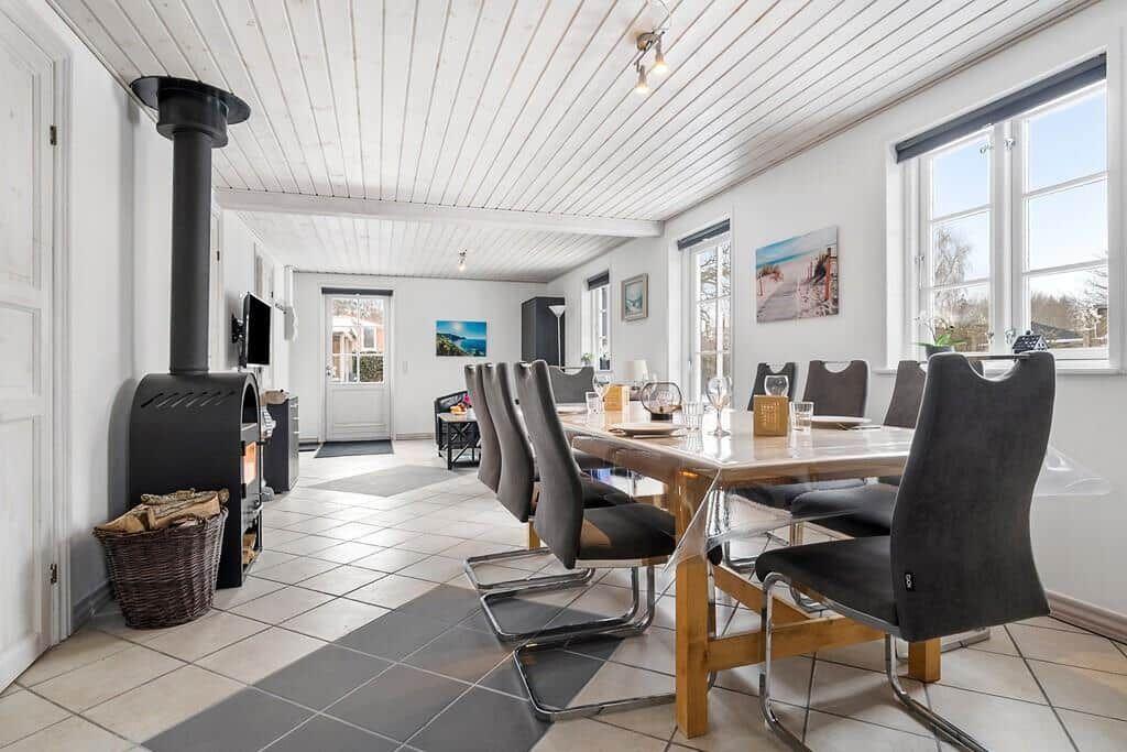 Dining room with wooden table, chairs, and wood stove. White walls and large windows.