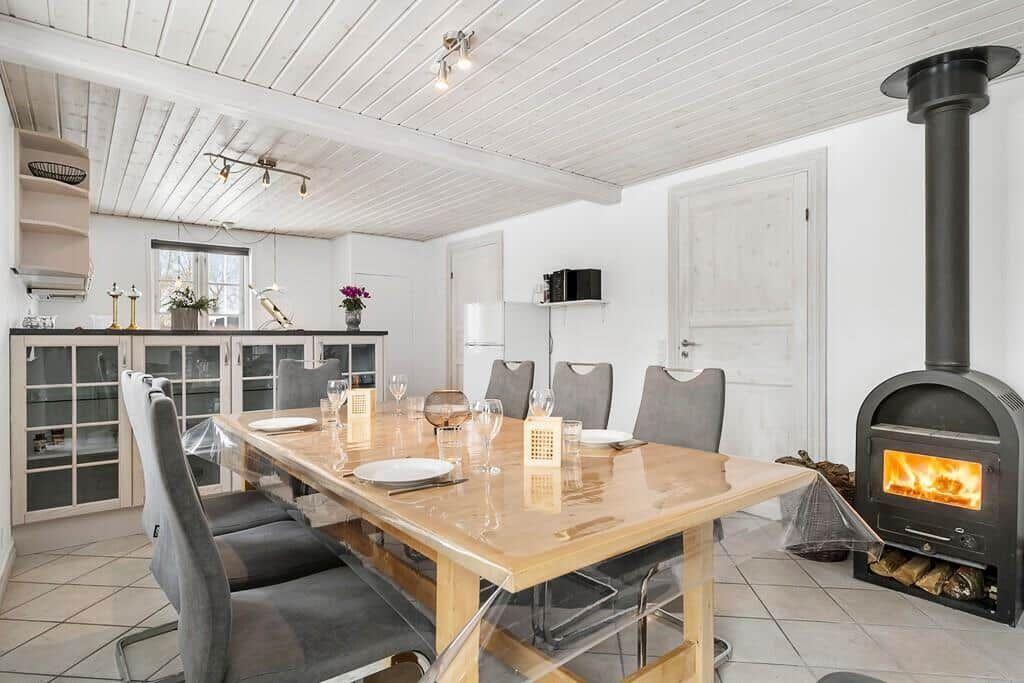Dining area with wooden table, chairs, and wood stove. White walls and ceiling.