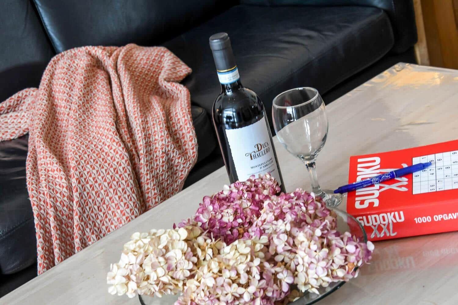 Auf dem Tisch steht eine Weinflasche, ein Glas, ein Sudoku und eine Vase mit Hortensien.