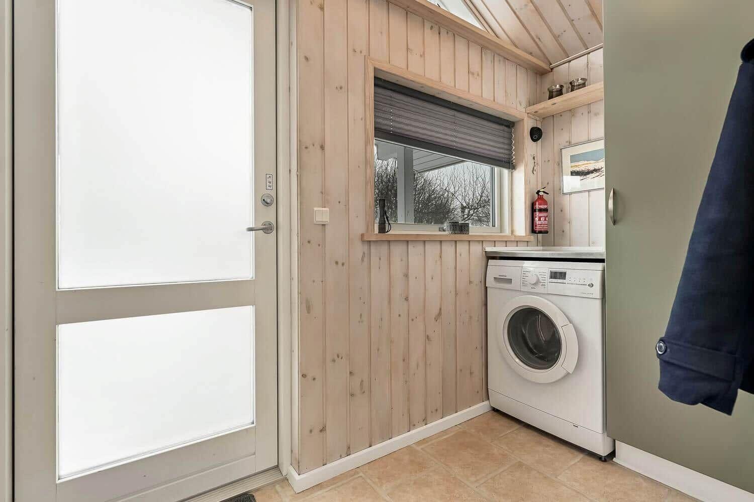 Waschmaschine in einem Raum mit Holzwand und Fenster. Tür und Kleidungsstück sind sichtbar.
