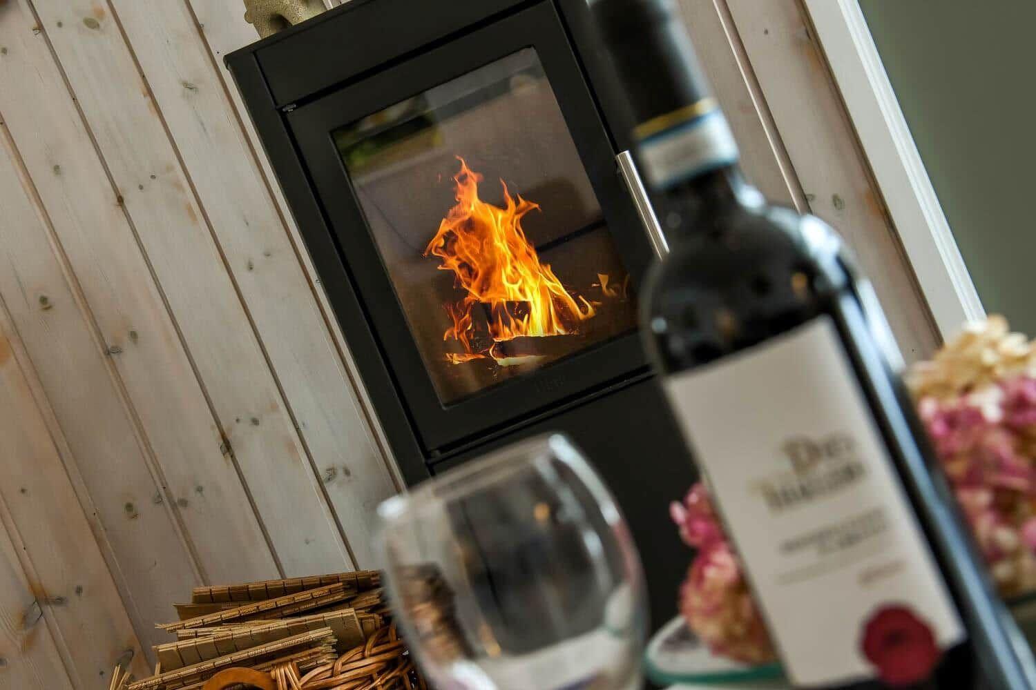 Kamin mit brennendem Feuer, Weinflasche und Glas auf Holzwand im Vordergrund.
