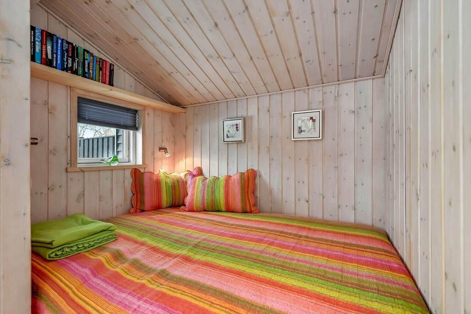 Schlafzimmer mit Holzwänden, Bett mit buntem Bettzeug und Fenster mit Jalousie.