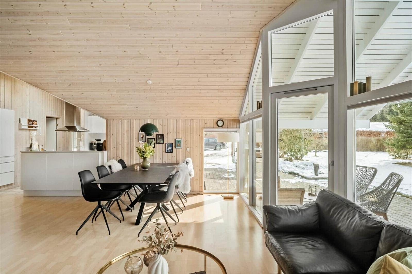 Large living area with dining space, kitchen, and large windows. Wooden ceiling and walls.