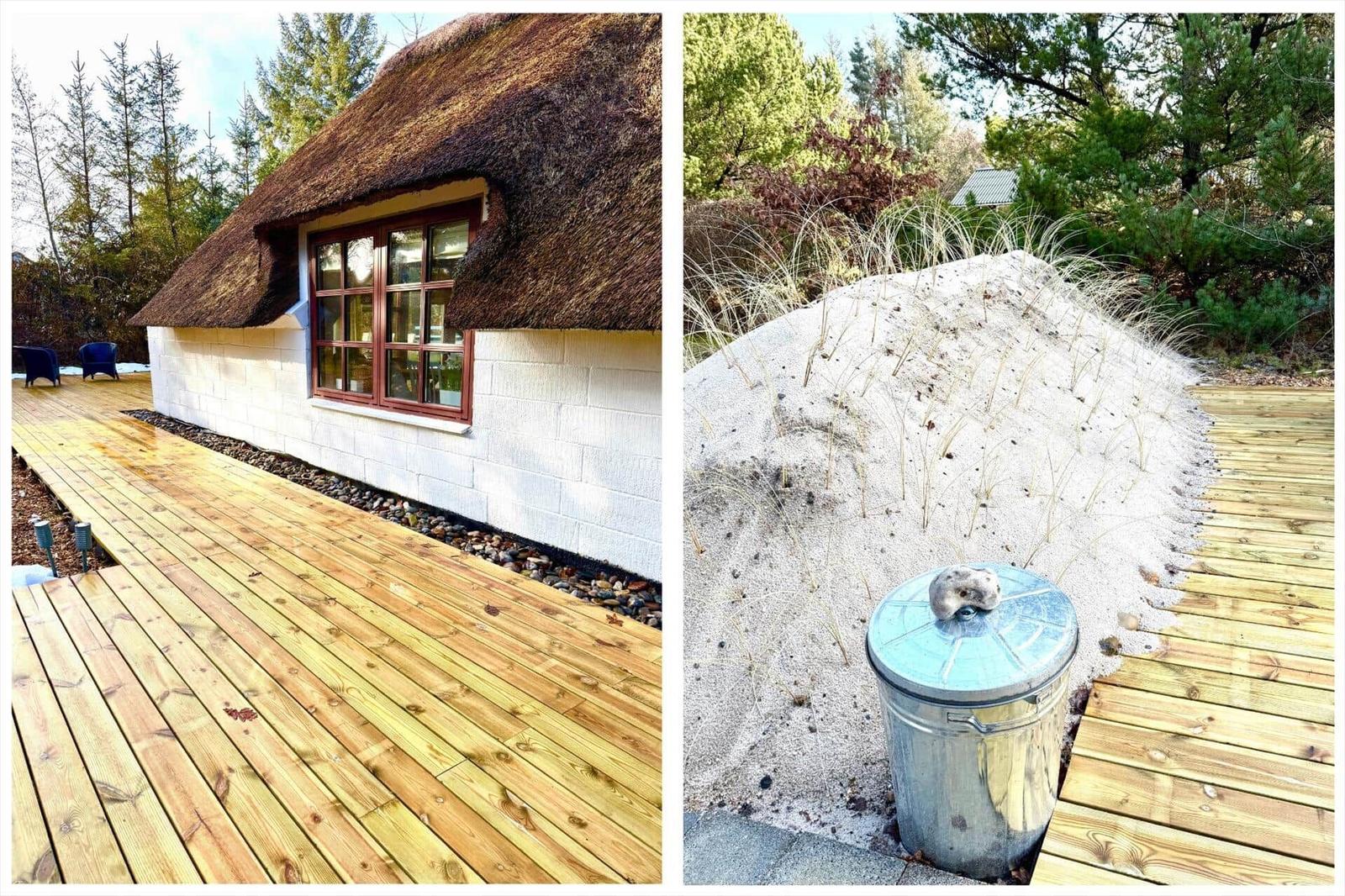 Haus mit Strohdach und Holzterrasse. Sandbeet mit Pflanzen und Metalltonne.
