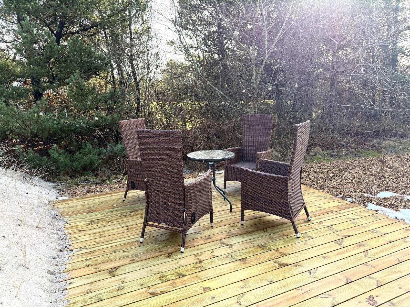 Terrasse mit vier Stühlen und Tisch, umgeben von Bäumen und Sand.