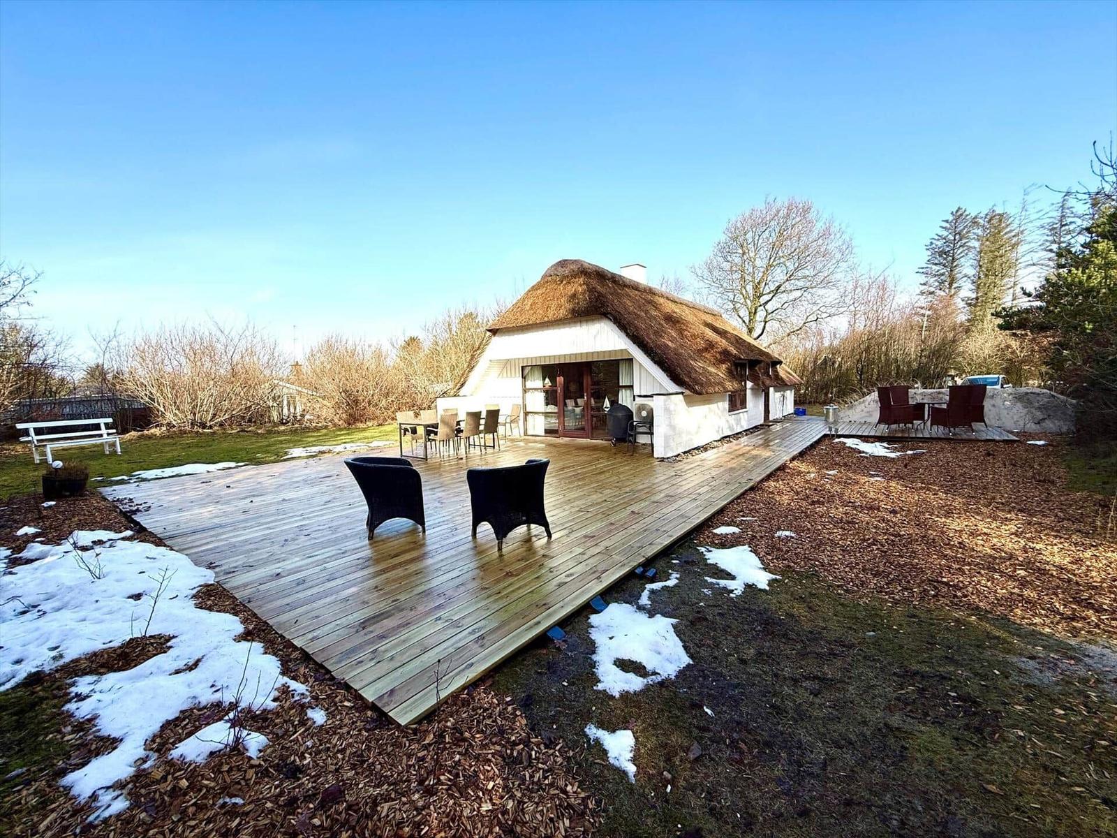 Haus mit strohgedecktem Dach und großer Holterrasse mit Sitzgelegenheiten.