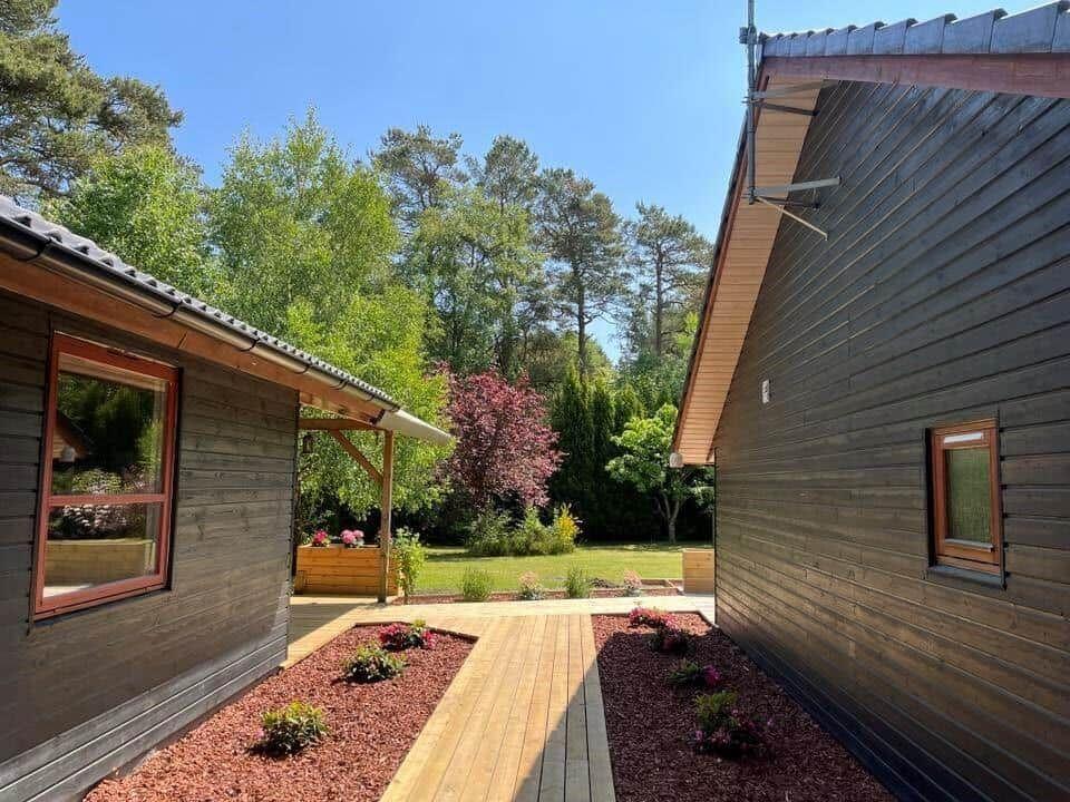 Holzhaus mit Terrasse und Garten, umgeben von Bäumen und Blumenbeeten.