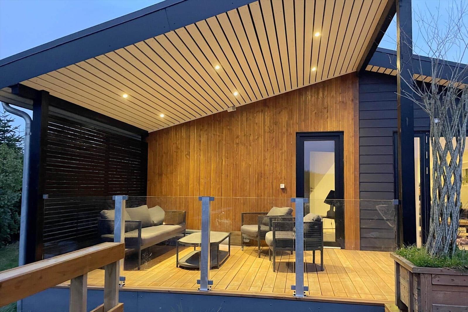 Terrasse mit Sitzgruppe und Holzwand. Glasgeländer und Deckenbeleuchtung.