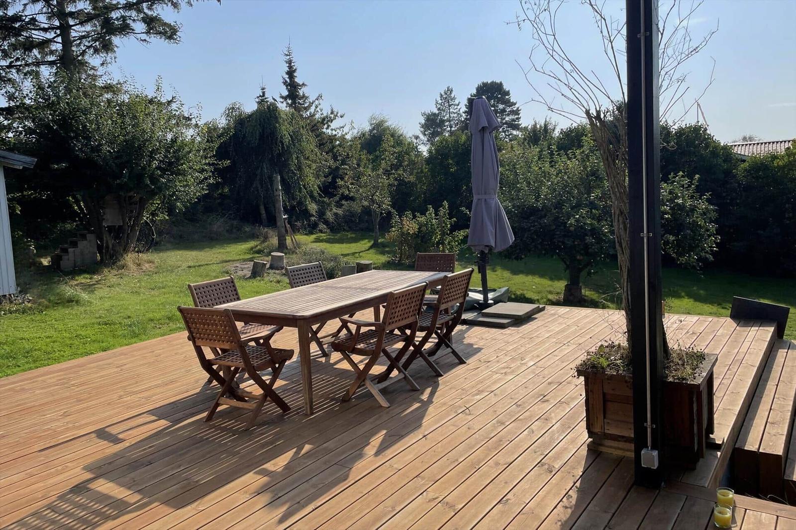 Holzterrasse mit Tisch, Stühlen und Sonnenschirm. Grünfläche und Bäume im Hintergrund.