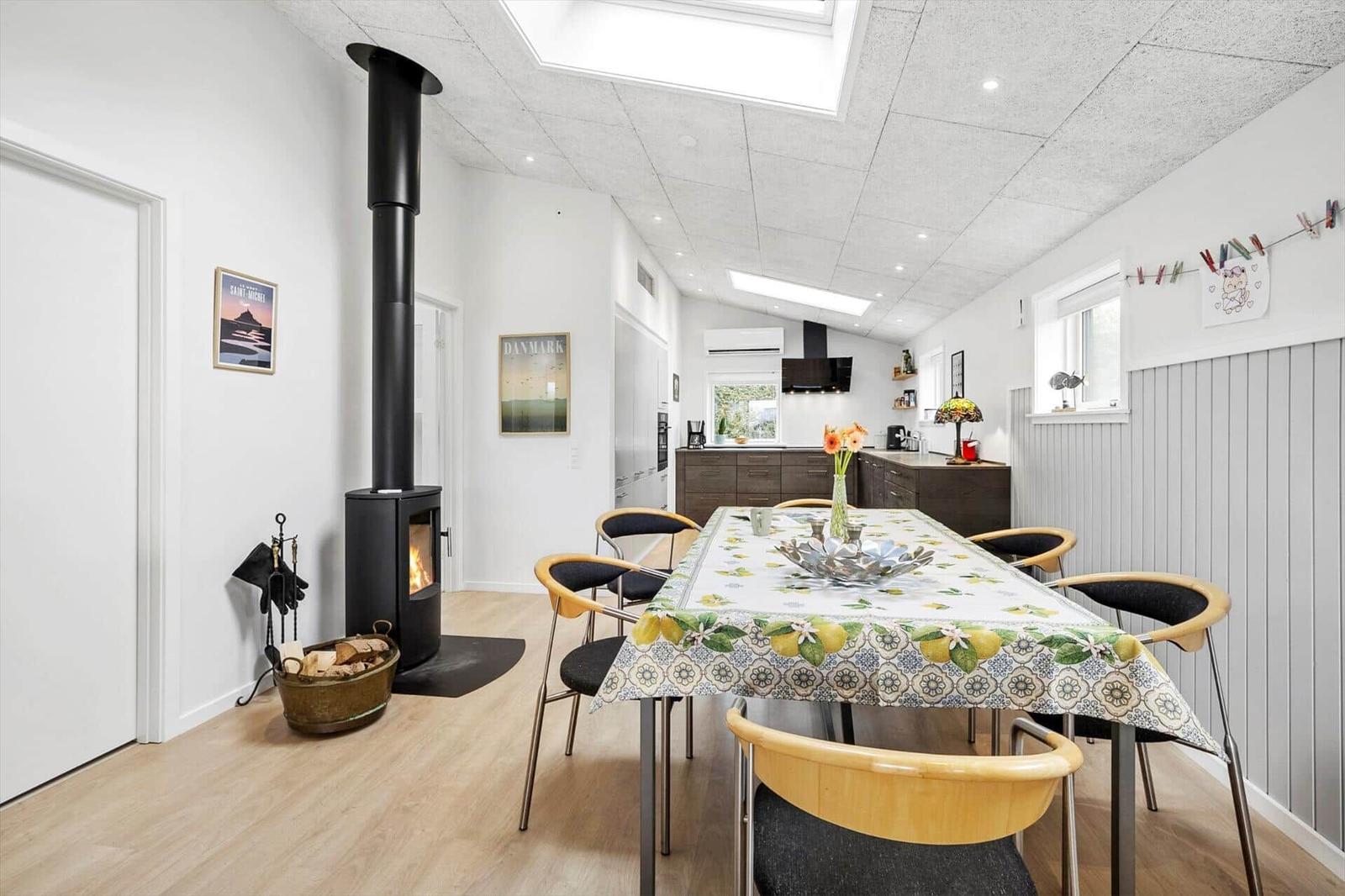 Esszimmer mit Tisch, Stühlen und Kaminofen. Küche im Hintergrund.