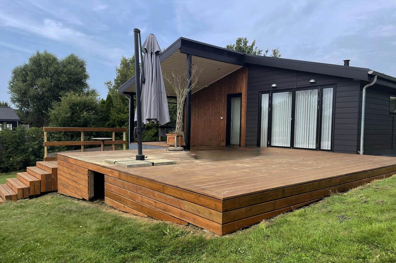 Moderne Villa mit Holzterrasse und Sonnenschirm. Grün umgeben.