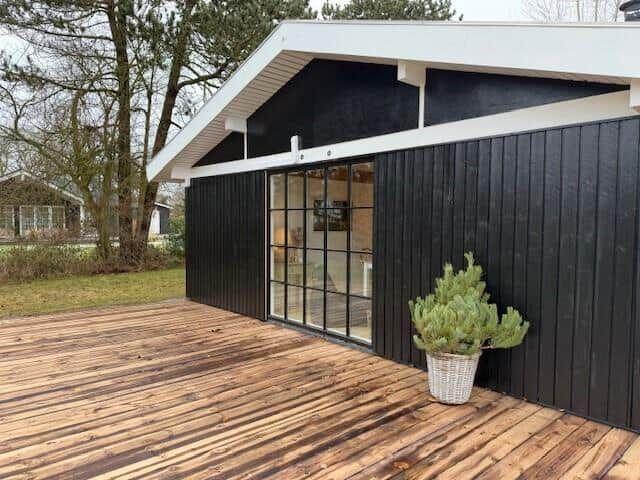 Moderne Hütte mit Holzterrasse und Glasfront. Pflanze im Korb steht vor der Wand.