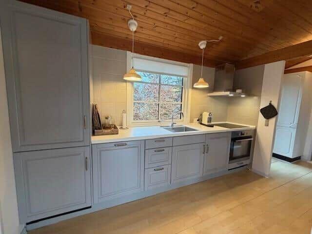 Küche mit hellen Schränken, Arbeitsplatte und Fenster. Holzdecke und Boden.