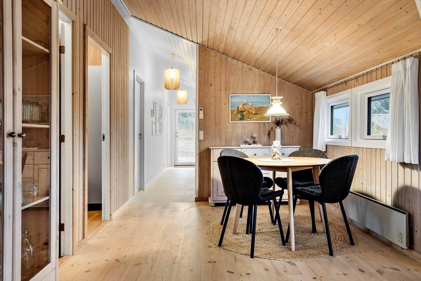 Dining area with round table and chairs, wooden walls and floor, windows with curtains.