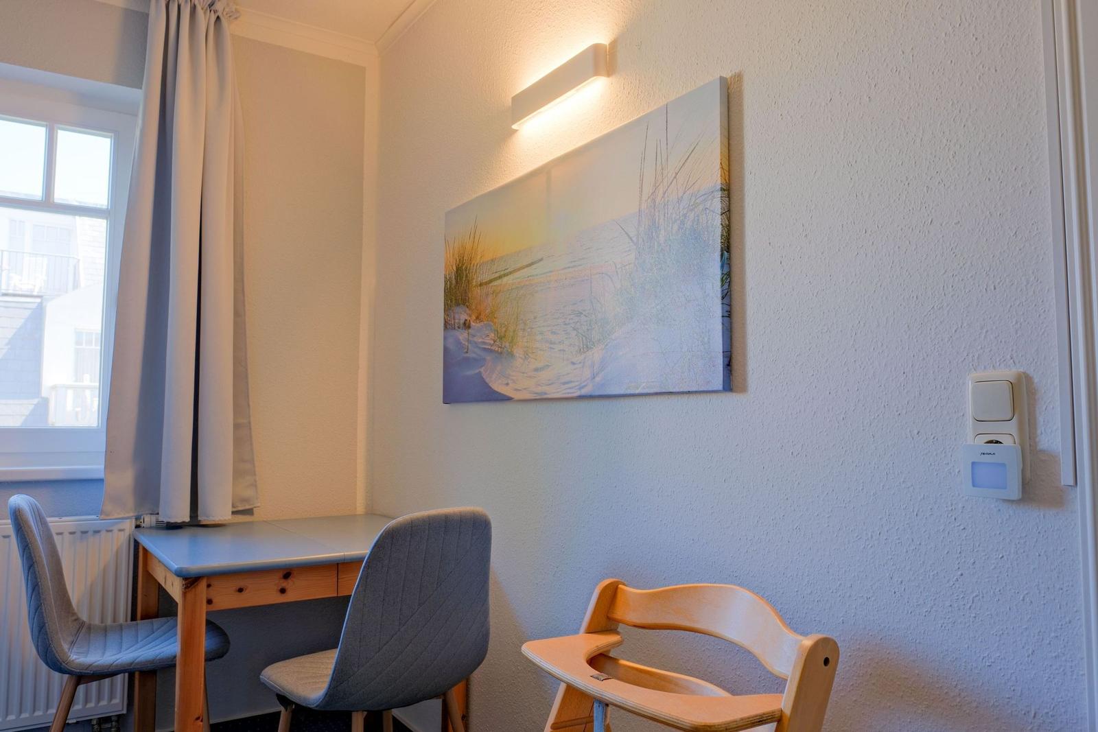 Schreibtisch mit zwei Stühlen und Kinderhochstuhl an Wand mit Strandbild.