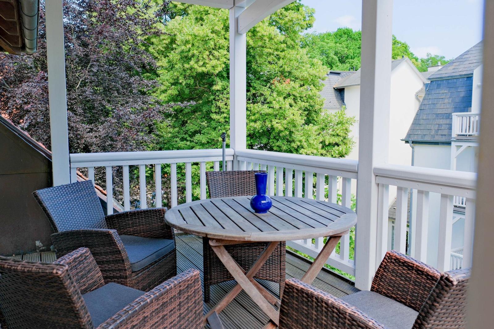 Terrasse mit runden Tisch und vier Stühlen, Blick auf grüne Bäume und benachbarte Häuser.