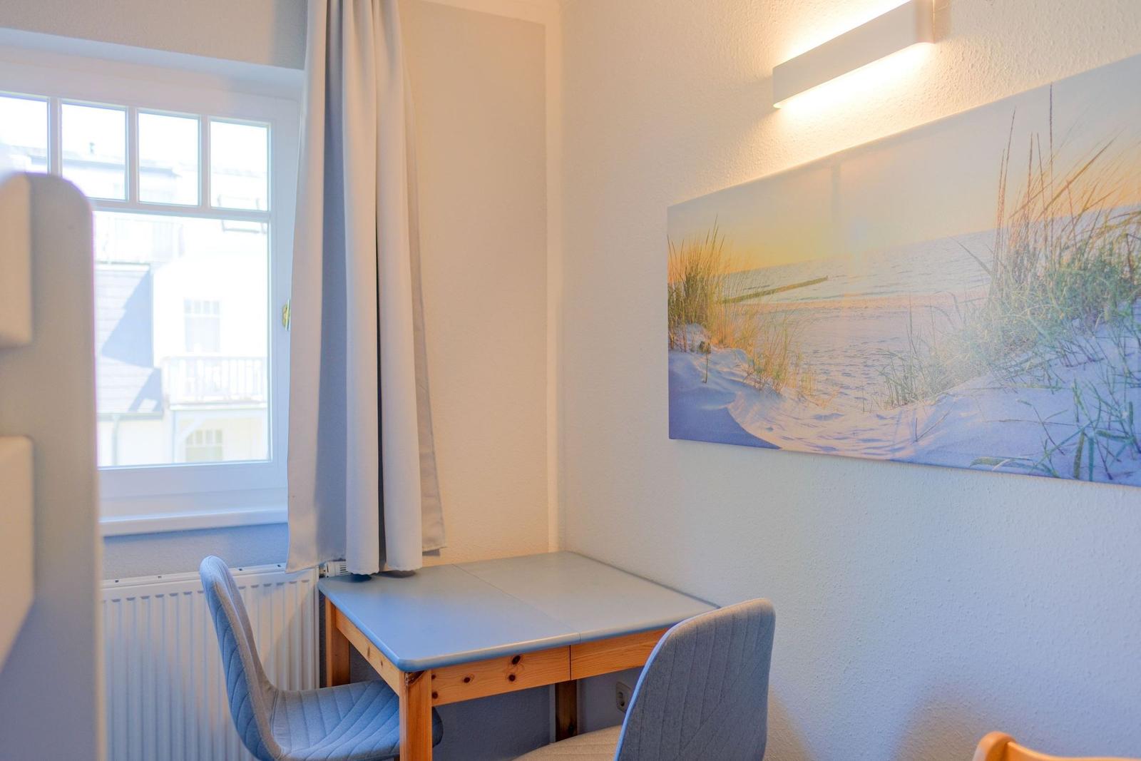 Ein Raum mit Tisch, zwei Stühlen und Wandbild. Fenster mit Vorhang. Heizung unter dem Fenster.