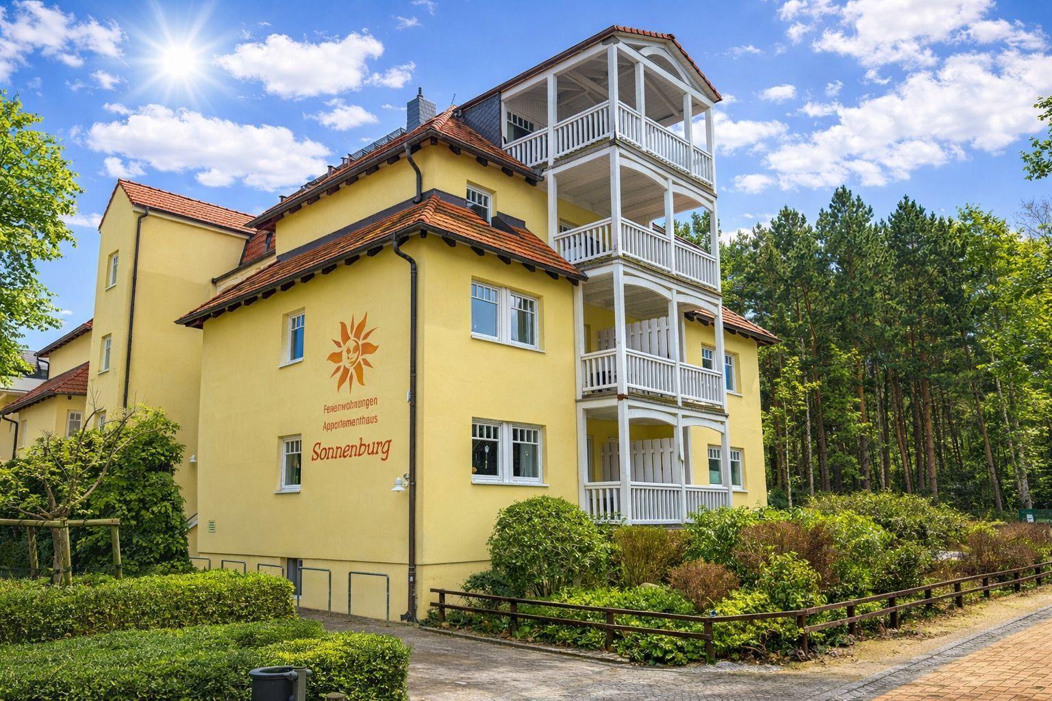 Gelbes Mehrfamilienhaus mit weißen Balkonen und rotem Dach. Umgeben von Bäumen und Grünflächen.