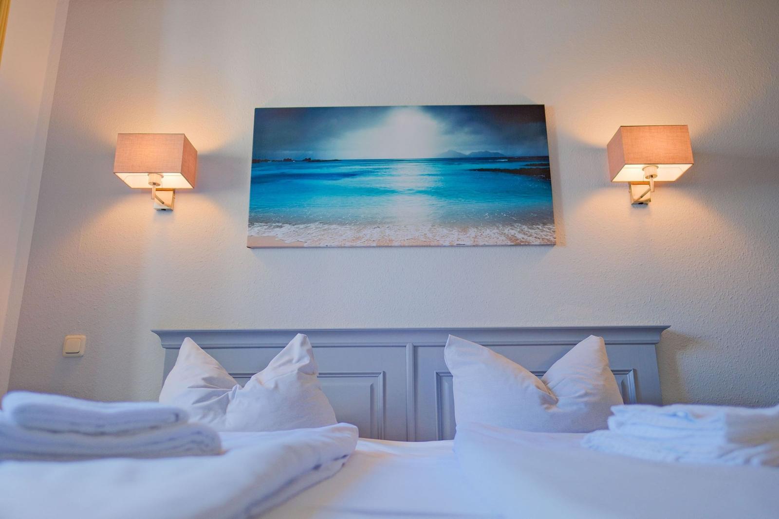 Ein Bett mit weißen Kissen und Handtüchern. Über dem Kopfteil hängt ein Strandbild mit zwei Wandlampen.