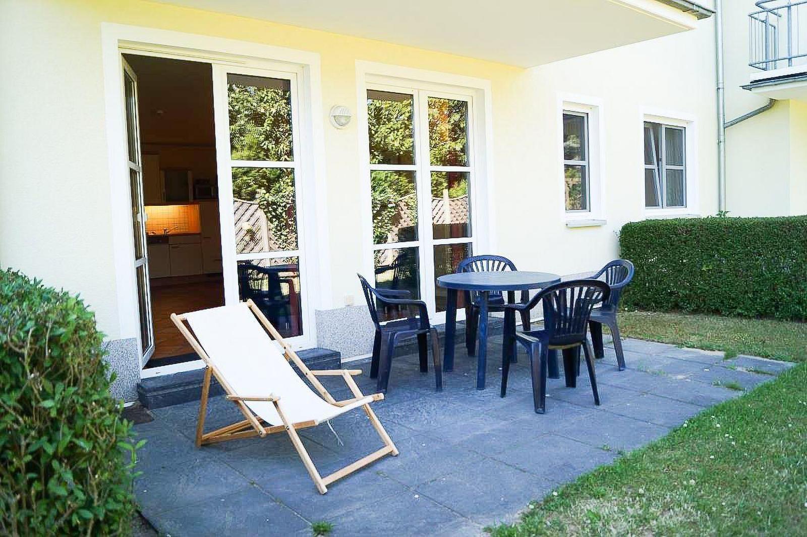 Terrasse mit Tisch, Stühlen und Liegestuhl. Durchgang ins Haus. Grünflächen umgeben.