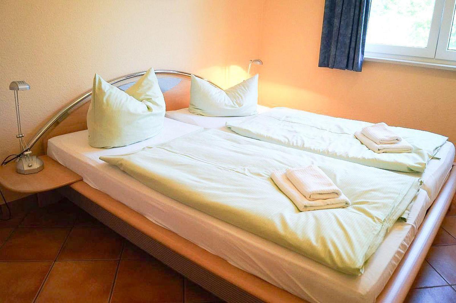 Doppelbett mit Bettwäsche und Handtüchern, Nachttisch mit Lampe, Fenster mit Vorhang.