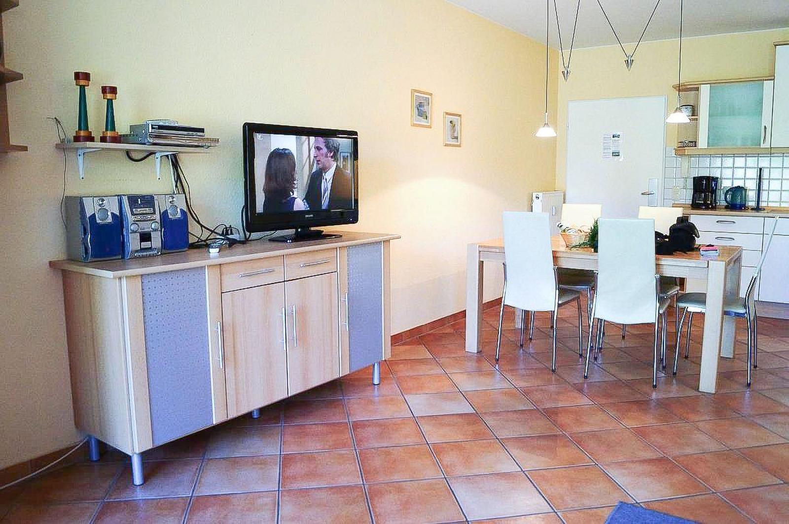 Wohn- und Essbereich mit TV, Tisch und Stühlen sowie Küche im Hintergrund.