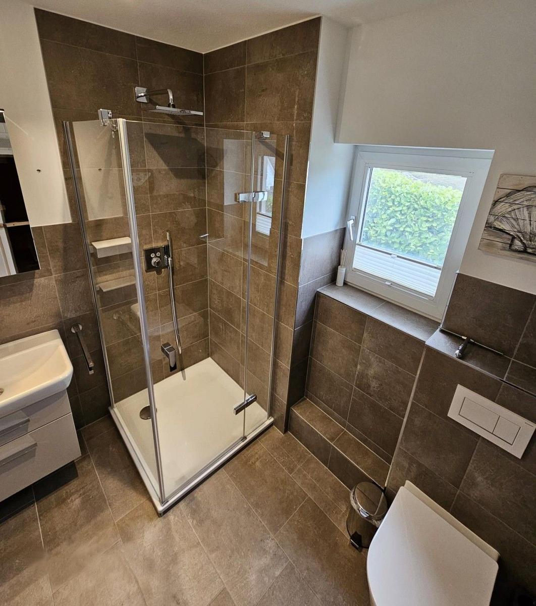 Badezimmer mit Dusche, Waschbecken und Toilette. Fenster mit Blick auf Grün.