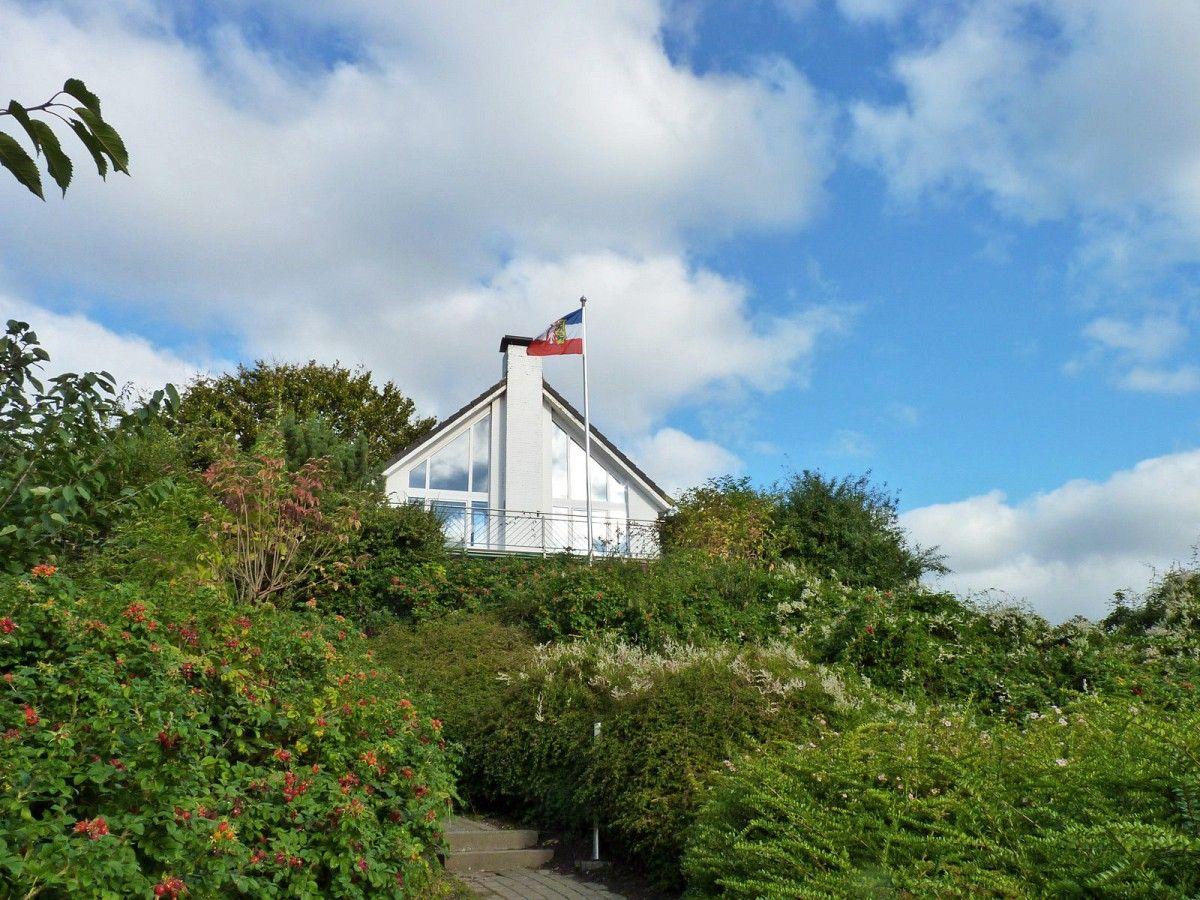 Weißes Haus mit Flagge auf Hügel, umgeben von grüner Vegetation und blauem Himmel.
